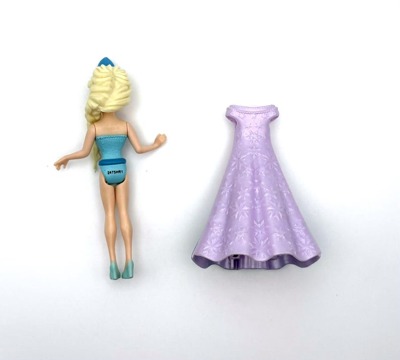 Pu&ograve; includere: Una bambola con i capelli biondi e un vestito blu &egrave; accanto a un vestito lilla chiaro. I capelli della bambola sono acconciati a treccia e l'abito ha un motivo floreale. La bambola indossa scarpe blu.
