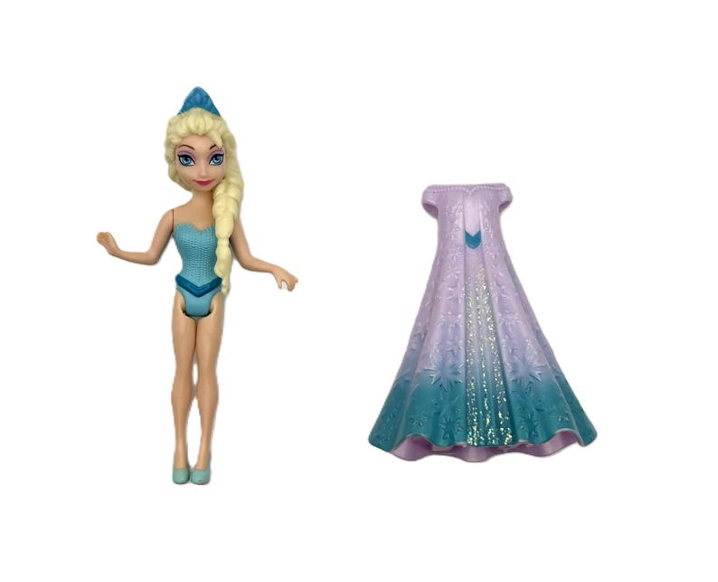 Pu&ograve; includere: Una bambola di un personaggio con i capelli biondi in una treccia laterale, che indossa un costume da bagno azzurro e una corona blu. Accanto alla bambola c'&egrave; un lungo mantello fluente nei toni del viola e del verde acqua.