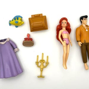 May include: A selection of miniature figurines and accessories. Includes a yellow and blue fish, a brown treasure chest, a red-haired doll in a pink bikini, a man in an orange jacket, a purple dress, a small red figure, and a gold candelabra.