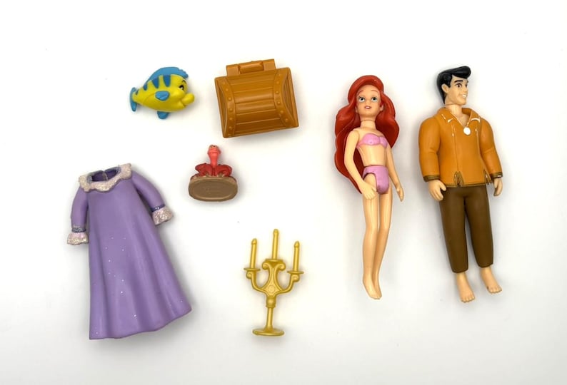 May include: A selection of miniature figurines and accessories. Includes a yellow and blue fish, a brown treasure chest, a red-haired doll in a pink bikini, a man in an orange jacket, a purple dress, a small red figure, and a gold candelabra.