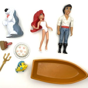 Op de afbeelding: Een verzameling speelgoed van Disney's De Kleine Zeemeermin, waaronder Ariel, Prins Eric, Botje, een boot, een hond, een taart, een drietand en een vork.