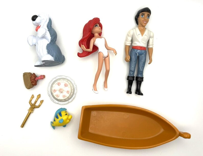 Op de afbeelding: Een verzameling speelgoed van Disney's De Kleine Zeemeermin, waaronder Ariel, Prins Eric, Botje, een boot, een hond, een taart, een drietand en een vork.