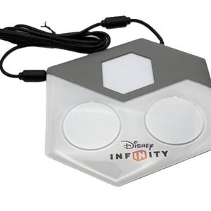 May include: A Disney Infinity base station with a gray and white design. The base has two circular areas and a hexagonal top section. The Disney Infinity logo is printed on the front. A black cable extends from the top.