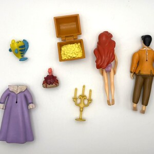 May include: A selection of plastic toy figurines and accessories. The set includes a yellow and blue fish, a treasure chest with gold coins, a red-haired mermaid, a man in an orange jacket, a purple robe, and a gold candelabra.