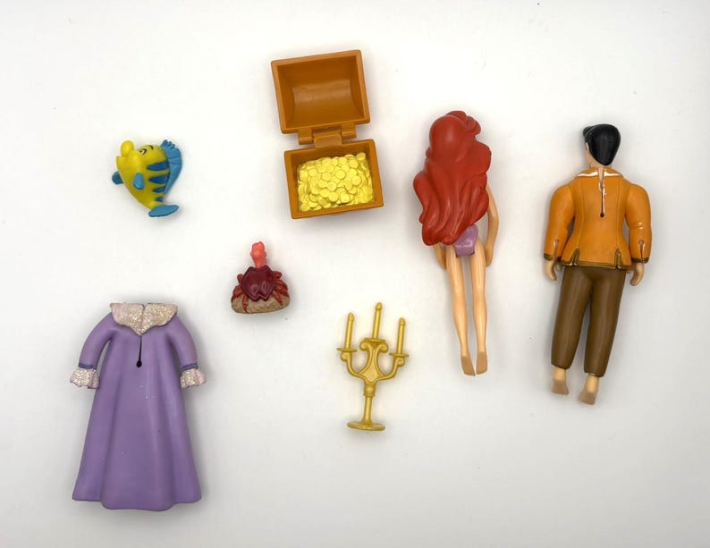 May include: A selection of plastic toy figurines and accessories. The set includes a yellow and blue fish, a treasure chest with gold coins, a red-haired mermaid, a man in an orange jacket, a purple robe, and a gold candelabra.