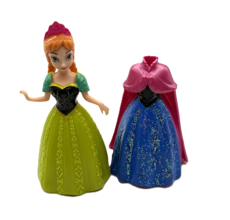 May include: Two plastic figurines of Anna from the movie Frozen. One figurine is wearing a green and yellow dress with a black bodice. The other figurine is wearing a blue dress with a pink cape.