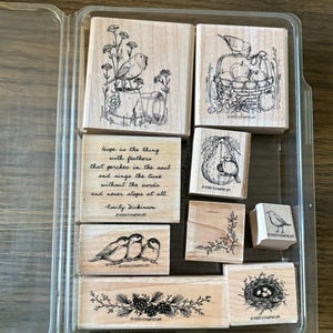 May include: A set of eight wooden rubber stamps in a clear plastic case. The stamps feature black ink illustrations of birds, a bird nest with eggs, a basket of apples, pine branches, and a quote by Emily Dickinson: Hope is the thing with feathers that perches in the soul and sings the tune without the words and never stops at all.