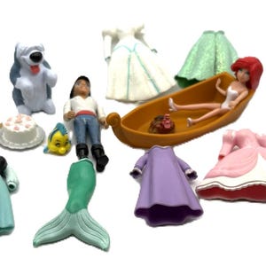 Op de afbeelding: Een verzameling Disney-beeldjes en accessoires, waaronder een pop in een boot, jurken, een zeemeerminstaart en karakterfiguren. De beeldjes zijn in verschillende kleuren, waaronder blauw, groen, roze en paars.