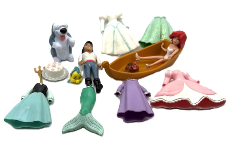 Op de afbeelding: Een verzameling Disney-beeldjes en accessoires, waaronder een pop in een boot, jurken, een zeemeerminstaart en karakterfiguren. De beeldjes zijn in verschillende kleuren, waaronder blauw, groen, roze en paars.