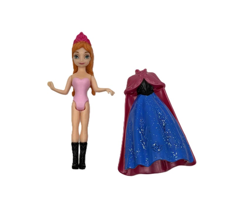 Pu&ograve; includere: Una bambola con i capelli rossi, un body rosa e stivali neri &egrave; in piedi accanto a un vestito blu e rosso. La bambola ha una corona rosa ed &egrave; in piedi. L'abito ha un mantello rosso.