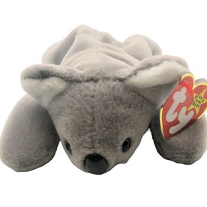 May include: A gray plush koala bear with a white belly and a black nose. The bear is lying on its belly with its paws outstretched. The bear has a red and white Ty tag attached to its right ear that says "Beanie Original Baby".
