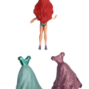 May include: A red-haired doll with a crown, wearing a teal swimsuit, and two dresses. One dress is teal and the other is purple with a floral pattern.