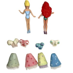 May include: Two small plastic dolls with blonde and red hair, wearing swimsuits. Several miniature doll accessories are also present, including tops and skirts in various colors like pink, blue, and green. The accessories have glitter and floral patterns.