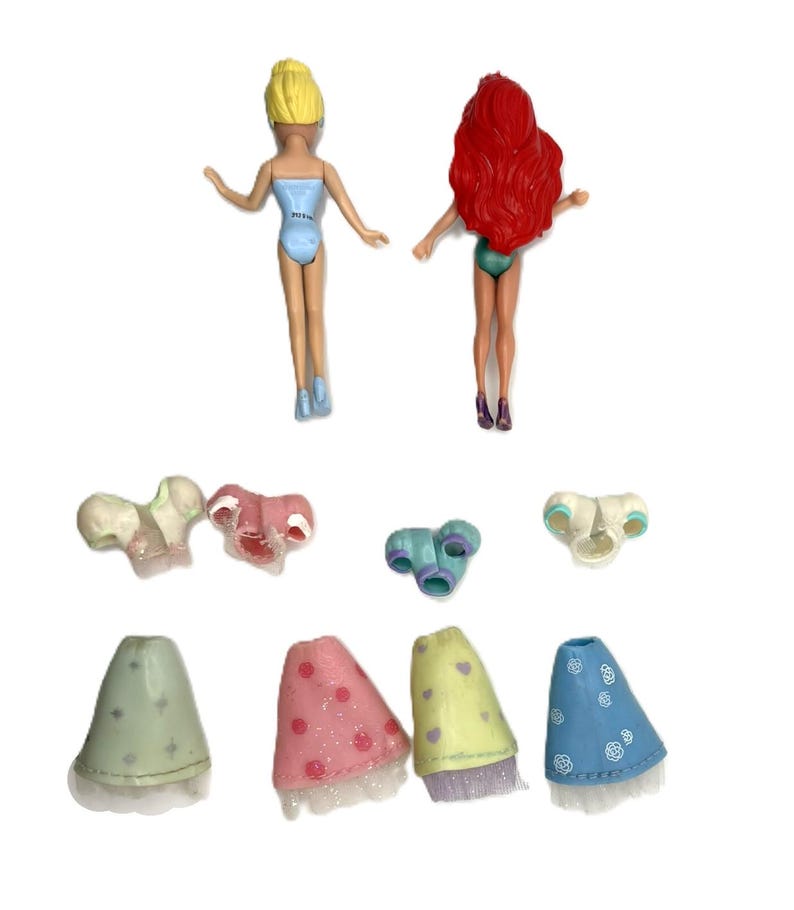 May include: Two small plastic dolls with blonde and red hair, wearing swimsuits. Several miniature doll accessories are also present, including tops and skirts in various colors like pink, blue, and green. The accessories have glitter and floral patterns.