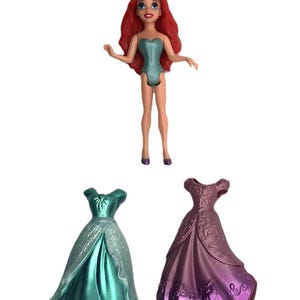 May include: A red-haired doll wearing a teal swimsuit and a silver crown. The doll is holding a teal dress and a purple dress.