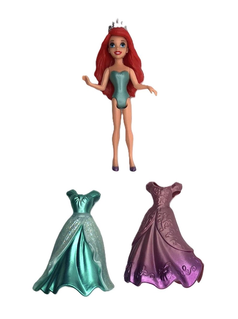 May include: A red-haired doll wearing a teal swimsuit and a silver crown. The doll is holding a teal dress and a purple dress.