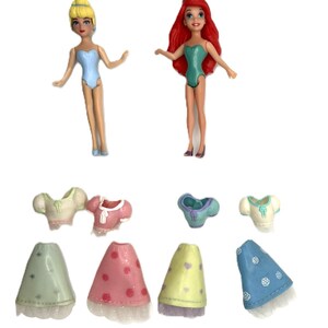 May include: Two small dolls of Disney princesses, Cinderella and Ariel, with removable outfits. Cinderella has blonde hair and a blue swimsuit. Ariel has red hair and a green swimsuit. The outfits include tops and skirts in various colors.