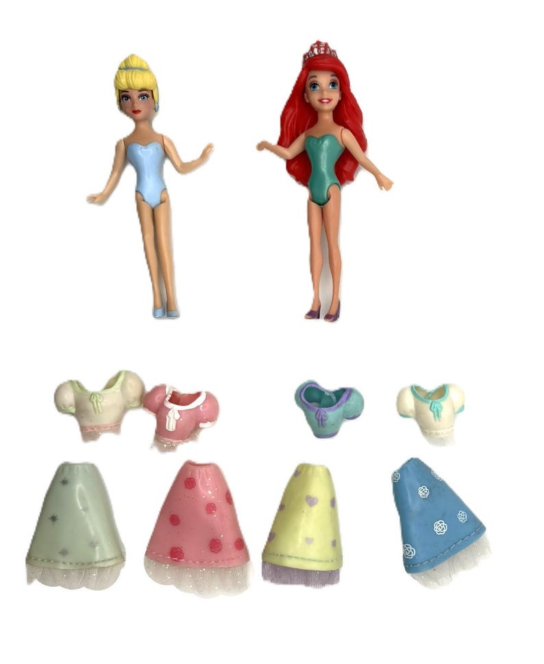 May include: Two small dolls of Disney princesses, Cinderella and Ariel, with removable outfits. Cinderella has blonde hair and a blue swimsuit. Ariel has red hair and a green swimsuit. The outfits include tops and skirts in various colors.