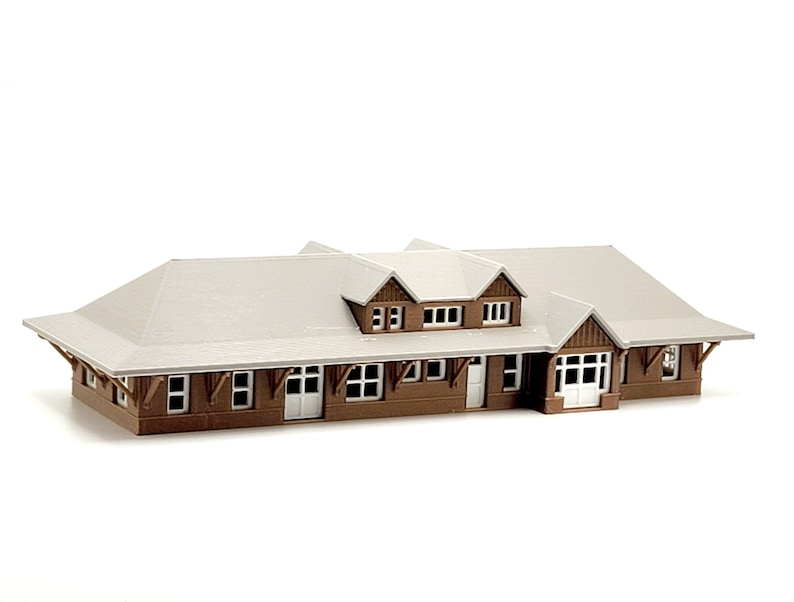 May include: A brown wooden model train station with a white roof and windows. The station has a front porch with a door and windows.