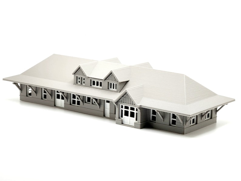 May include: A gray model train station with a gabled roof and multiple windows. The station has a covered platform and a small entrance.