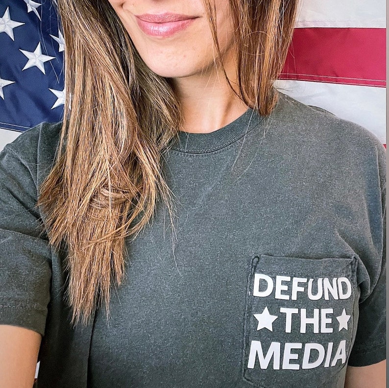 May include: A dark green t-shirt with a white pocket that says "DEFUND THE MEDIA" with a star on either side of the word "THE".