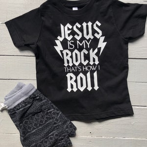 May include: Black t-shirt with white text that reads "Jesus is my rock that's how I roll". The text is in a rock and roll font style. The shirt is paired with black and white patterned shorts.