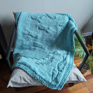 Peut inclure: Une couverture turquoise tricotée avec un motif texturé, drapée sur un coussin gris sur une chaise. La couverture présente un motif répété de poissons stylisés en relief. La chaise a un cadre en métal foncé.