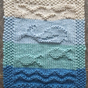 Op de afbeelding: Handgebreid vierkant met een zeelandschapontwerp. Het bovenste gedeelte is gebroken wit, gevolgd door lichtblauwe, blauwgroene en diepblauwe secties. Het ontwerp bevat getextureerde golven, wolken en vissen.
