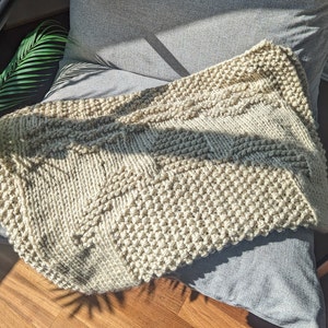 Peut inclure: Une couverture en tricot beige avec un motif texturé. La couverture est pliée et drapée sur une chaise.