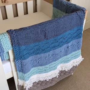 May include: A knitted baby blanket in shades of blue, green, and brown with a white fringe. The blanket is draped over a white crib.