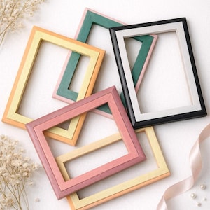 Puede incluir: Una colección de marcos de fotos rectangulares de colores rosa, amarillo, verde y negro. Los marcos están dispuestos sobre una superficie blanca con elementos decorativos, como flores secas y una cinta de satén.
