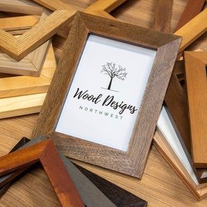 Puede incluir: Una colección de marcos de fotos de madera en varios tonos de marrón y beige. Un marco muestra el logotipo "Wood Designs Northwest" con un gráfico de árbol. Los marcos están dispuestos sobre una superficie de madera.