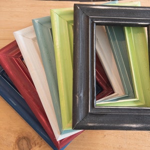 May include: A set of six picture frames in various colors, including black, green, blue, red, white, and gray. The frames are stacked on top of each other, with the black frame on top.