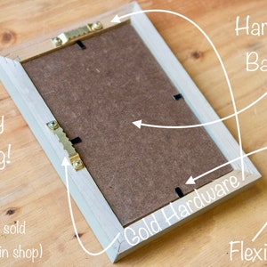 May include: The back of a light wood picture frame with a brown cardboard backing. The frame has gold-colored hanging hardware and flexi-tabs for easy hanging. The text "Ready to hang!" and "(Stands sold separately in shop)" is written on the frame.