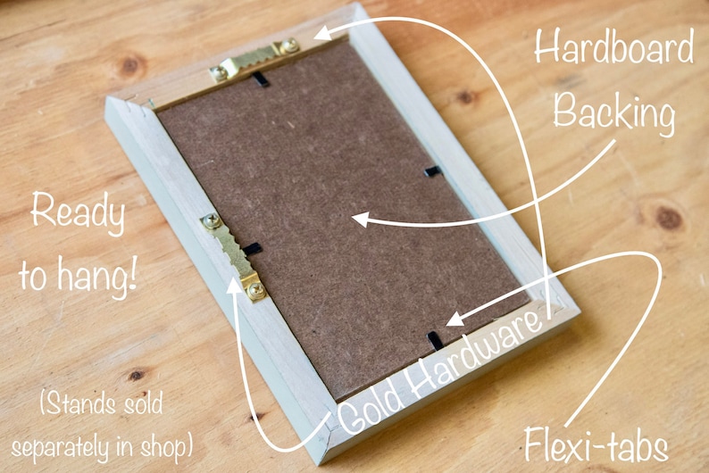 May include: The back of a light wood picture frame with a brown cardboard backing. The frame has gold-colored hanging hardware and flexi-tabs for easy hanging. The text "Ready to hang!" and "(Stands sold separately in shop)" is written on the frame.