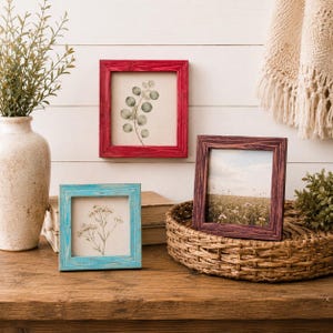 May include: Three square wooden picture frames in red, blue, and brown, each with a different nature-themed print. The frames are displayed on a wooden surface with a woven basket and a decorative vase.