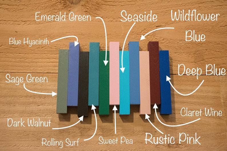 May include: A set of ten paint samples, each a different color, arranged in a row on a wooden surface. The colors are: Sage Green, Blue Hyacinth, Dark Walnut, Rolling Surf, Emerald Green, Sweet Pea, Seaside, Rustic Pink, Wildflower Blue, and Deep Blue.