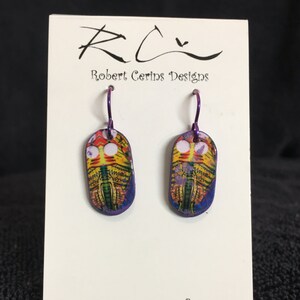 Könnte beinhalten: Ein Paar farbenfrohe, abstrakte Kunstohrringe mit lila Ohrhaken. Die Ohrringe sind oval geformt und haben ein lebendiges Design mit gelben, orangen, grünen und violetten Farbtönen. Auf der Karte steht "Robert Cerins Designs" und "Art In Miniature To Wear."