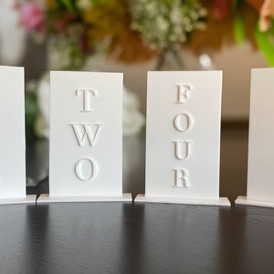 May include: Four white table number cards with embossed lettering spelling out "ONE", "TWO", "FOUR", and "THREE". The cards are rectangular and stand upright on small, attached bases. The background is blurred, with flowers visible.