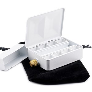 May include: A white metal paint box with a hinged lid and a brass knob. The box has nine compartments and is sitting on a black velvet pouch.