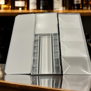 May include: A white metal paint palette with multiple compartments for holding paint. The palette is open and has a hinged lid.