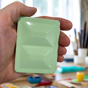 May include: A rectangular, light green watercolor palette held in a hand. The palette has a geometric design on the lid and appears to be empty. The background includes art supplies and a window.