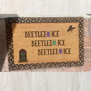 May include: A rectangular coir doormat with a brown border and a light brown center. The mat features the text "BEETLEJUICE" repeated three times, with a purple and green beetle graphic, and a tombstone that reads "NEVER TRUST THE LIVING". A black insect graphic is also present.