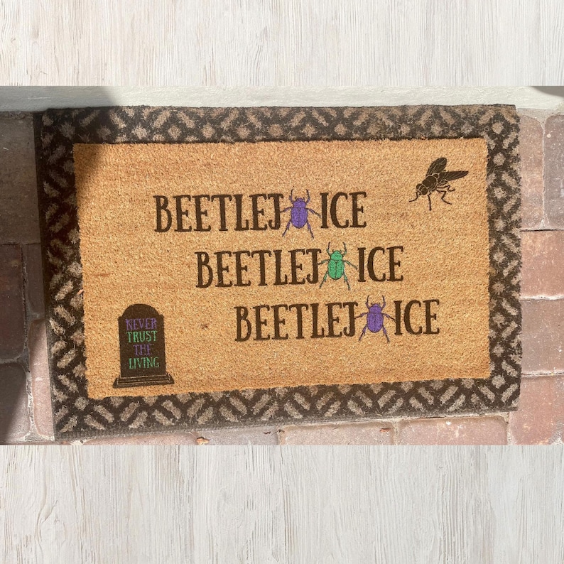 May include: A rectangular coir doormat with a brown border and a light brown center. The mat features the text "BEETLEJUICE" repeated three times, with a purple and green beetle graphic, and a tombstone that reads "NEVER TRUST THE LIVING". A black insect graphic is also present.
