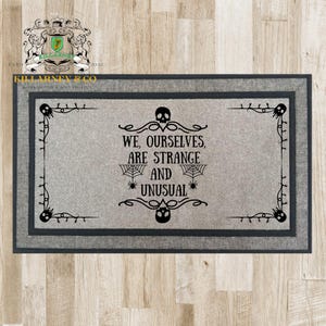 May include: Rectangular gray doormat with a dark gray border. The mat features the text "WE, OURSELVES, ARE STRANGE AND UNUSUAL" in black, along with spiderwebs, spiders, and skull designs. The doormat is a home decor item.