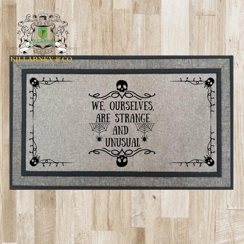 May include: Rectangular gray doormat with a dark gray border. The mat features the text "WE, OURSELVES, ARE STRANGE AND UNUSUAL" in black, along with spiderwebs, spiders, and skull designs. The doormat is a home decor item.