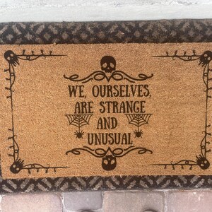 May include: Rectangular doormat with a brown coir center and a dark brown patterned border. The center features the text "WE, OURSELVES, ARE STRANGE AND UNUSUAL" in dark brown, with spiderwebs and skulls as design elements.