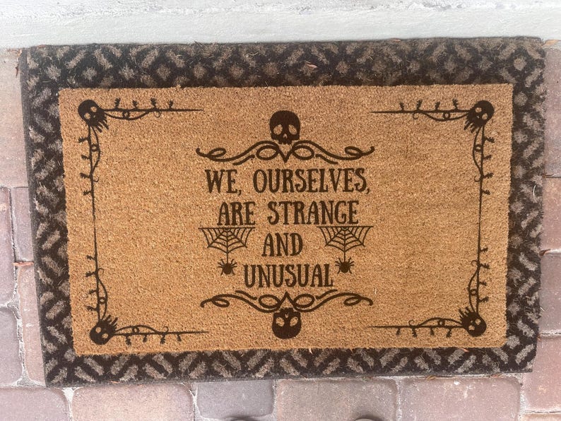 May include: Rectangular doormat with a brown coir center and a dark brown patterned border. The center features the text "WE, OURSELVES, ARE STRANGE AND UNUSUAL" in dark brown, with spiderwebs and skulls as design elements.
