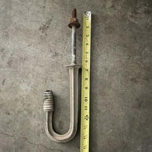 May include: A J-shaped metal hook with a threaded top section, a nut, and a ribbed end, positioned next to a yellow measuring tape that indicates a total length of approximately 15 inches from the top of the bolt to the bottom of the hook.
