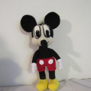 May include: A crocheted plush toy of Mickey Mouse. The toy is black with red shorts and yellow shoes. The toy has large black ears and a white face with black eyes.